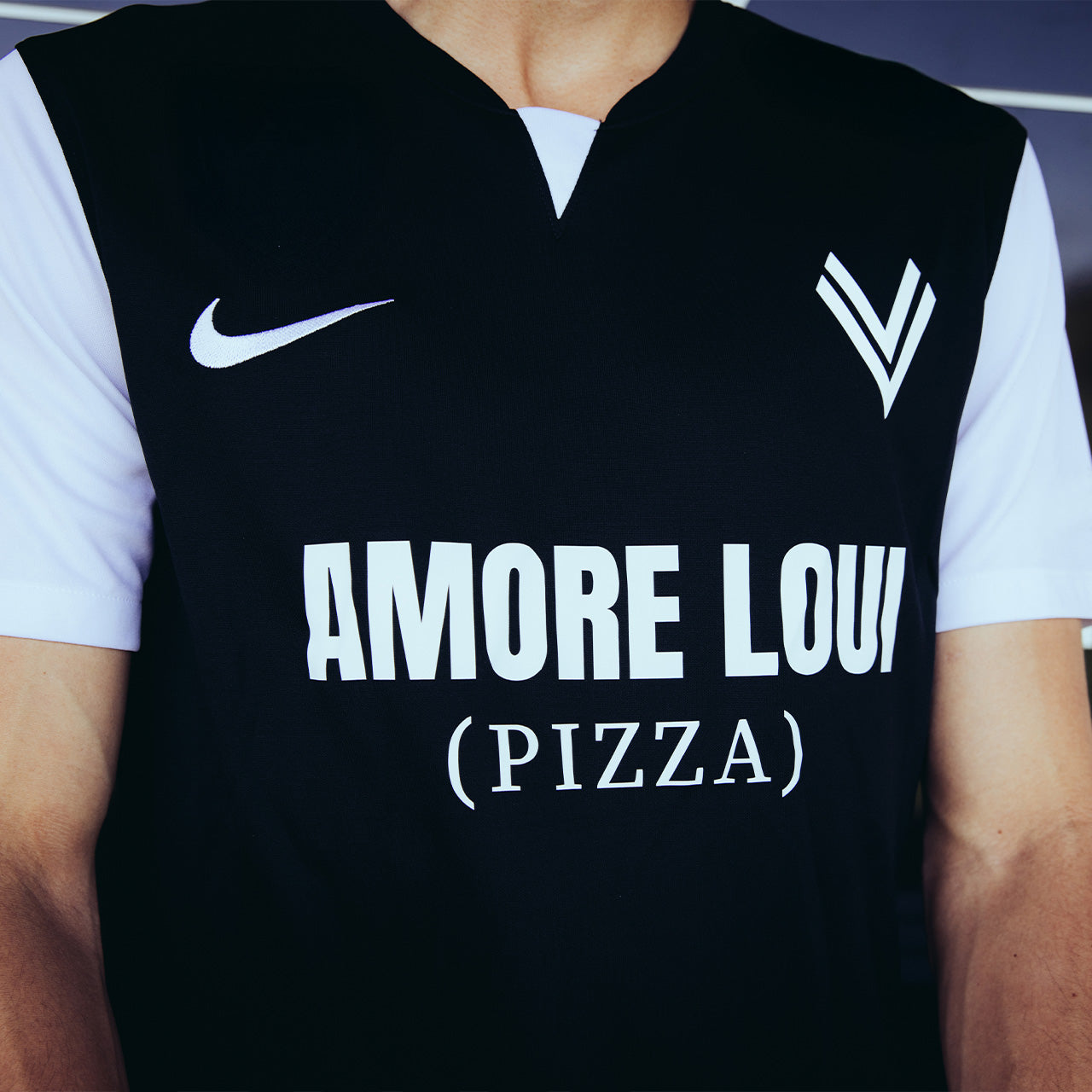 Mann trägt schwarzes ASK Voitsberg Trikot mit weißem kurzen Ärmeln mit weißem ASK Voitsberg Wappen auf der linken Brustseite, weißem Nike Swoosh auf der rechten Brustseite und Amore Loui (Pizza) Logo in weiß über die gesamte Brust.