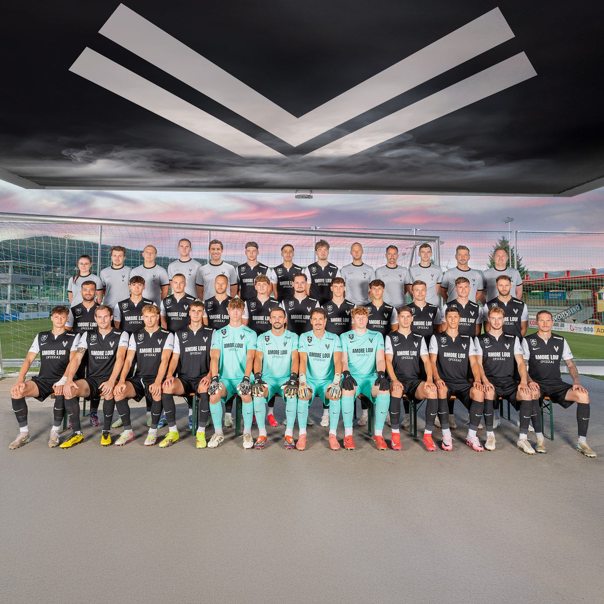 ASK Voitsberg Kader 25/26 Gruppenbild am Sportplatz bei Sonnenuntergang mit Fußball Tor im Hintergrund.