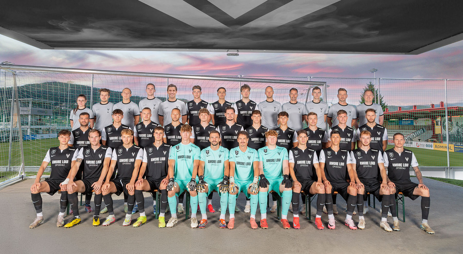 ASK Voitsberg Kader 25/26 Gruppenbild am Sportplatz bei Sonnenuntergang mit Fußball Tor im Hintergrund.