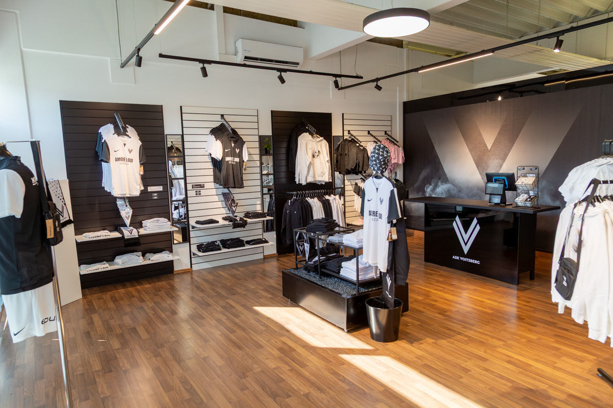 Moderner ASK Voitsberg Sportartikel-Laden mit Kleidung, Taschen und Accessoires, die ordentlich in Regalen und auf Kleiderständern präsentiert werden, mit Holzboden und einer schwarzen Theke mit einer Kasse in der hinteren Ecke.