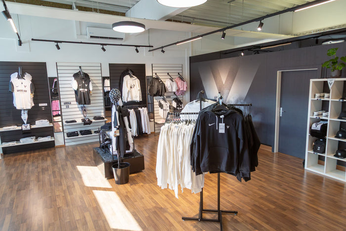 Moderner Sportartikel-Laden mit Kleidung, Taschen und Accessoires, die ordentlich in Regalen und auf Kleiderständern präsentiert werden, mit Holzboden und einer schwarzen Theke mit einer Kasse in der hinteren Ecke.