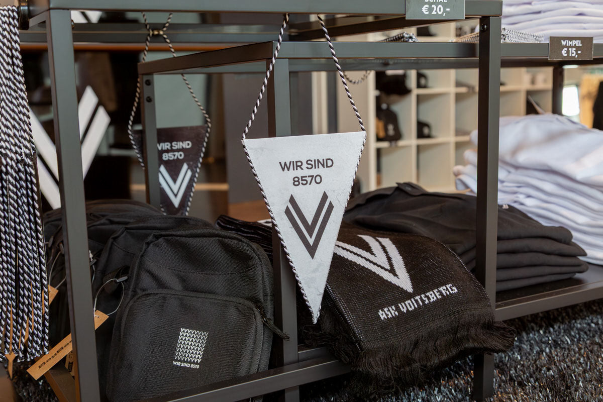 Verkaufsbereich direkt bei Eingang des ASK Voitsberg Teamshops in Voitsberg auf welchem mehrere Produkte wie Fanwimpel, Fanschals und Polos für den Verkauf präsentiert werden.