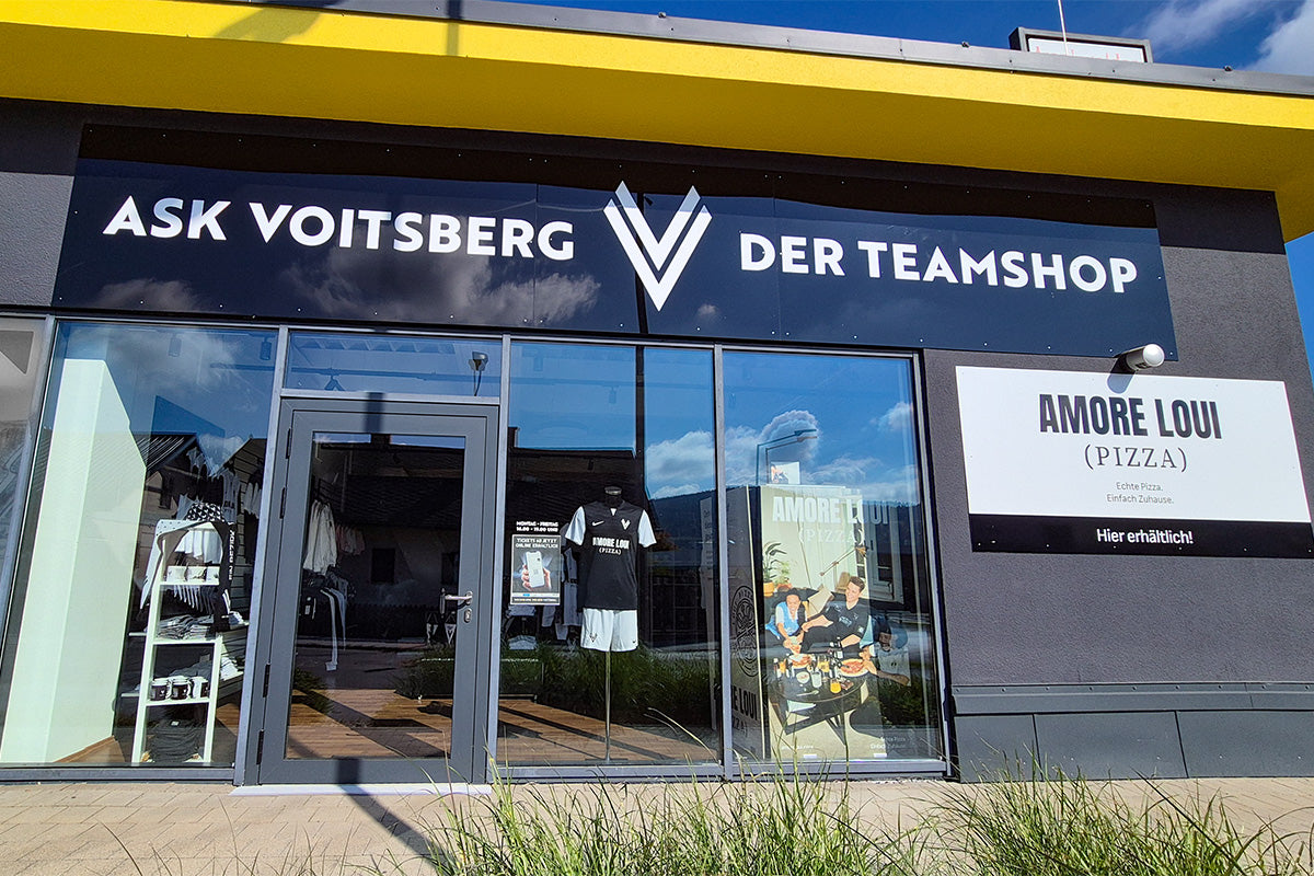 Dunkelgraue Fassade und gelbem Dachrand, Schriftzug 'ASK VOITSBERG DER TEAMSHOP' und Schild 'AMORE LOUI (PIZZA)' an der Wand. Ansicht der Präsentation Glasfassade.