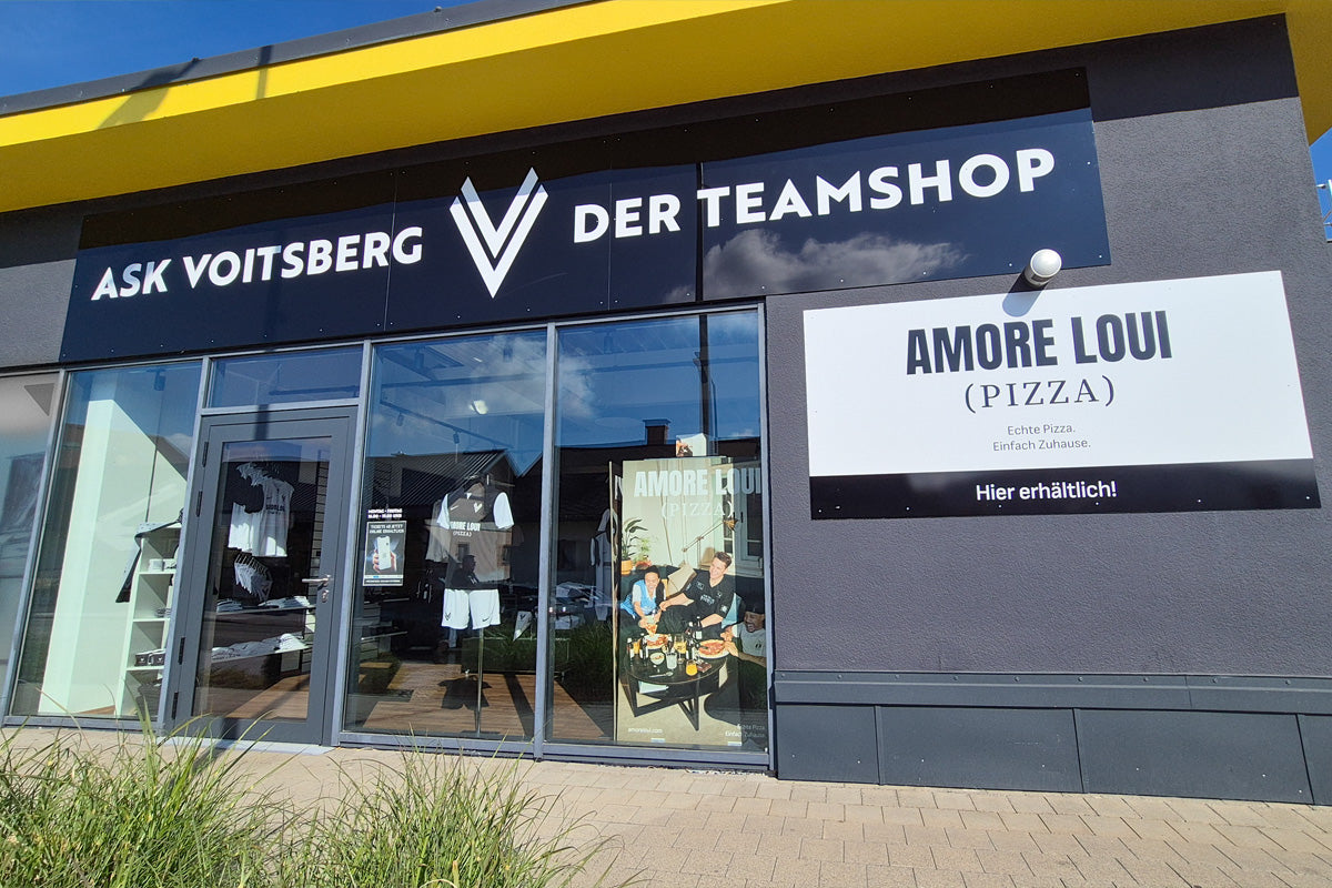 Dunkelgraue Fassade und gelbem Dachrand, Schriftzug 'ASK VOITSBERG DER TEAMSHOP' und Schild 'AMORE LOUI (PIZZA)' an der Wand. Ansicht der Präsentation Glasfassade.
