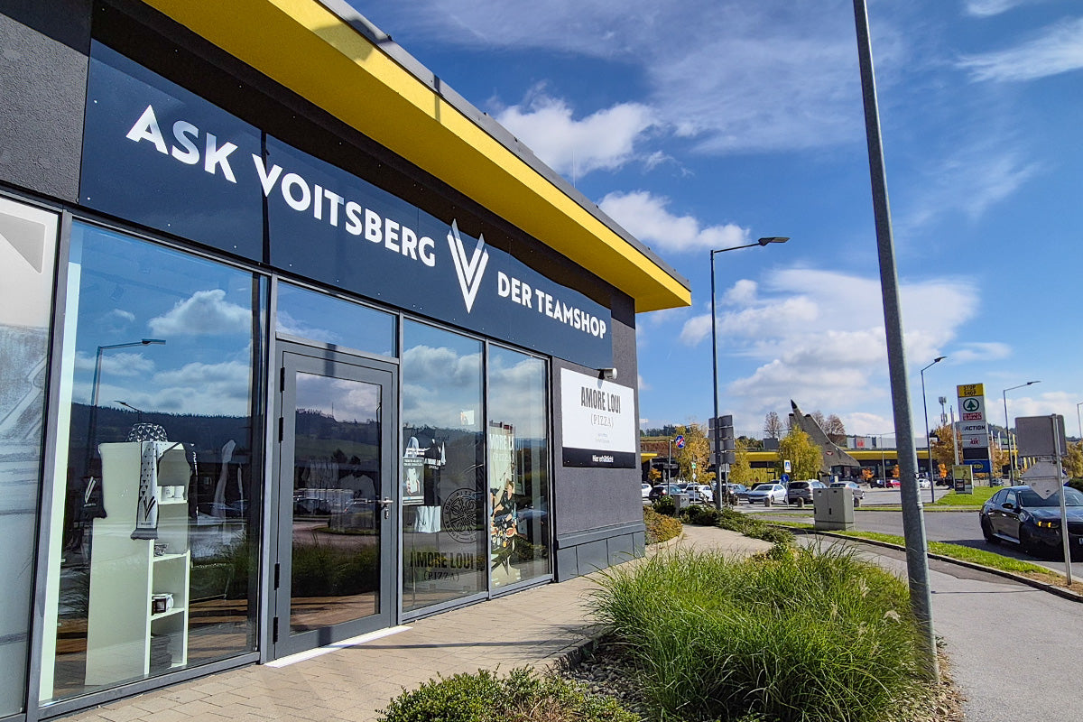 Dunkelgraue Fassade und gelbem Dachrand, Schriftzug 'ASK VOITSBERG DER TEAMSHOP' und Schild 'AMORE LOUI (PIZZA)' an der Wand. Ansicht der Präsentation Glasfassade. Im Hintergrund sieht man das den Parkplatz des Shoppingcenters.