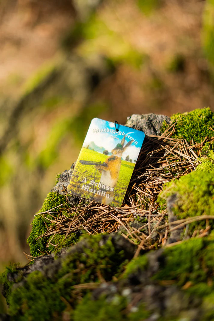 Duftbam 'A Schuss Marülln' Vorderseite auf bemoostem Stein. MJV Logo in weiß und Hase mit Schrottflinte in den Pfoten. 'A Schuss Marülln' Duftbaum vom Marderjagdverein.