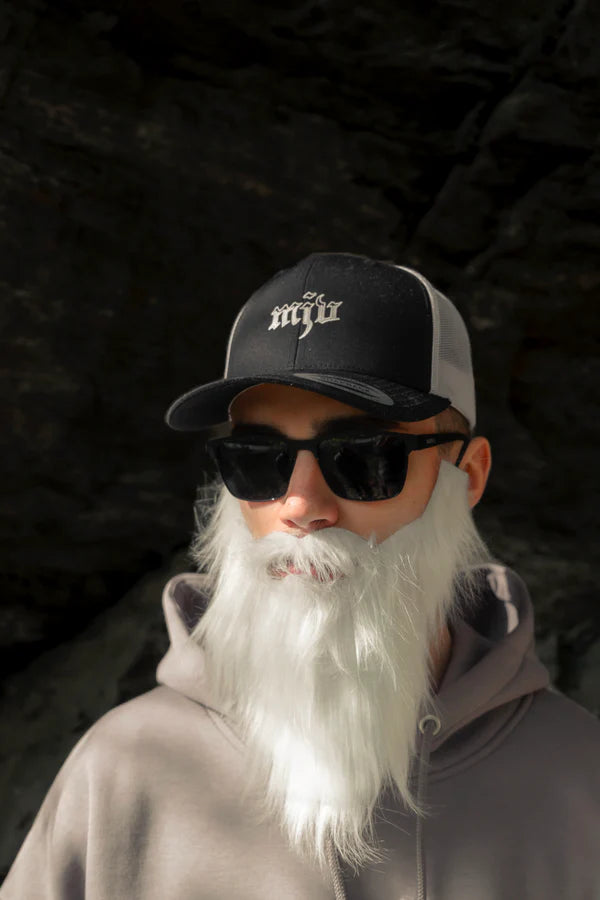 Mann trägt einen langen weißen Bart, Sonnenbrille und ein schwarz-weißes Trucker-Cap mit gesticktem weißen 'MJV' Logo.
