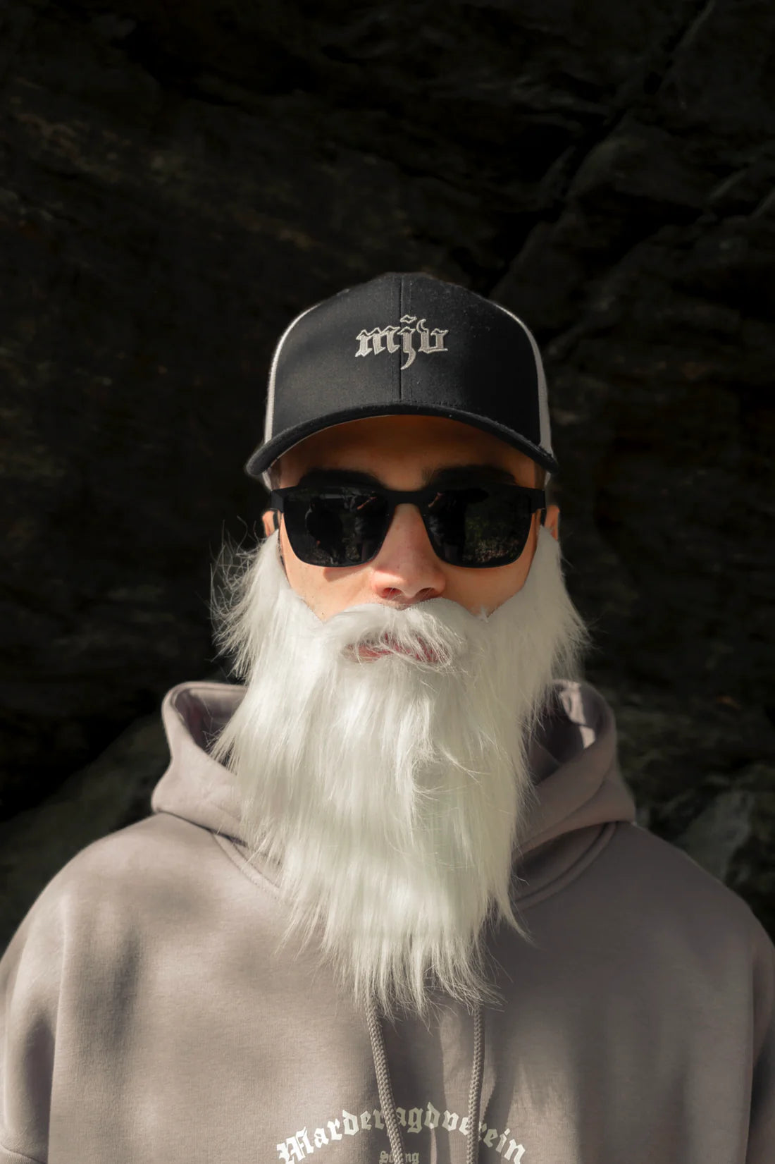 Mann trägt einen langen weißen Bart, Sonnenbrille und ein schwarz-weißes Trucker-Cap mit gesticktem weißen 'MJV' Logo sowie einen grauen Kapuzenpullover mit weißem Schriftzug auf der Brust.