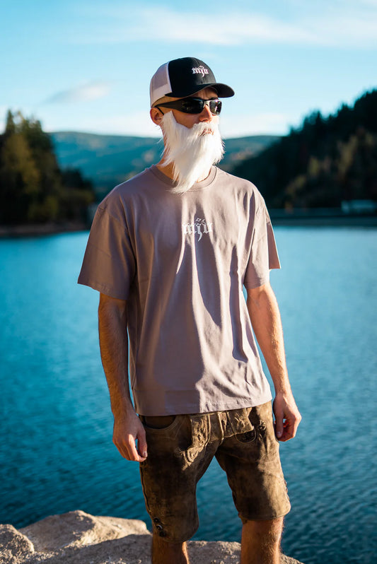 Ein Mann steht vor einem blauen See und Bergen. Er trägt eine schwarz-weiße Kappe mit Logo, eine Lederhose und ein graues Kurzarm-T-Shirt mit weißem Logo.