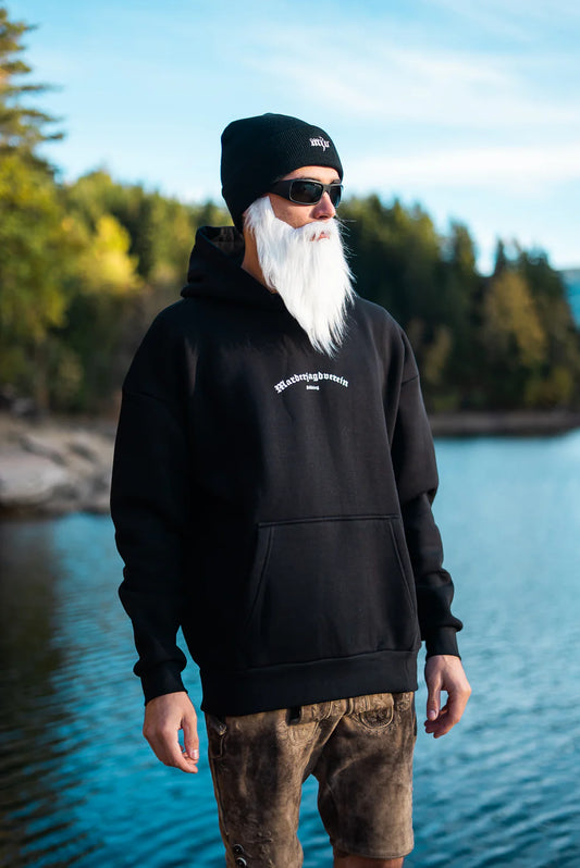 Ein Mann steht vor einem blauen See und Bergen. Er trägt einen weißen langen Bart und einen schwarzen Kapuzenpullover mit weißer 'Marderjagdverein Söding' Aufschrift.